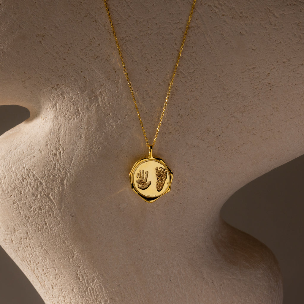 The Baby Hand & Footprint Necklace in 18K Gold features a round pendant engraved with your baby's hand and footprint, displayed on a textured surface—an ideal personalized keepsake and baby jewelry piece.