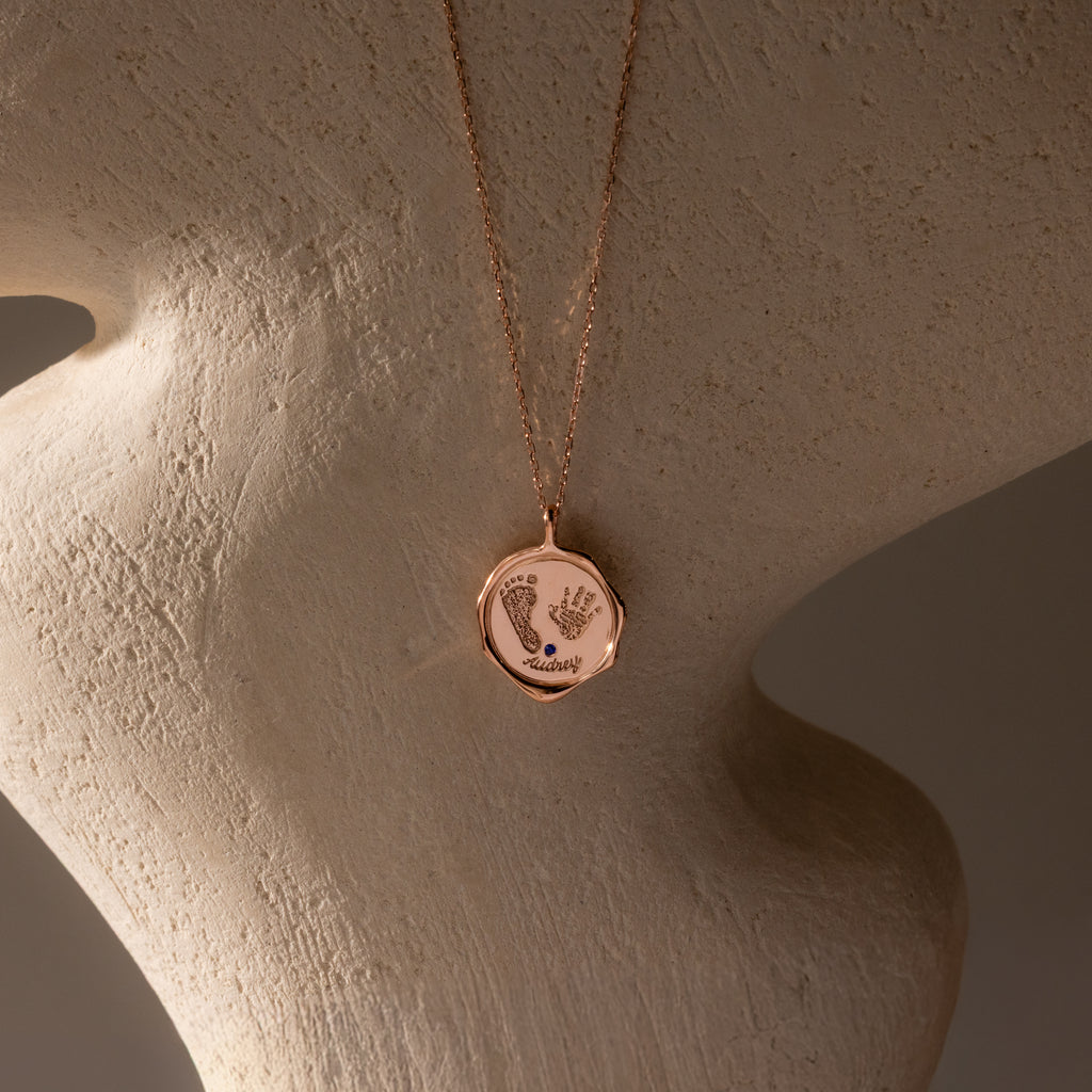 The Baby Hand & Footprint Necklace in Rose Gold features a pendant engraved with delicate hand and footprints, displayed on a textured beige bust—a heartfelt personalized jewelry keepsake.