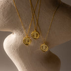 The Baby Hand & Footprint Necklace in 18K Gold, along with two other engraved gold keepsake necklaces, are displayed on a textured, curved, neutral-colored surface.