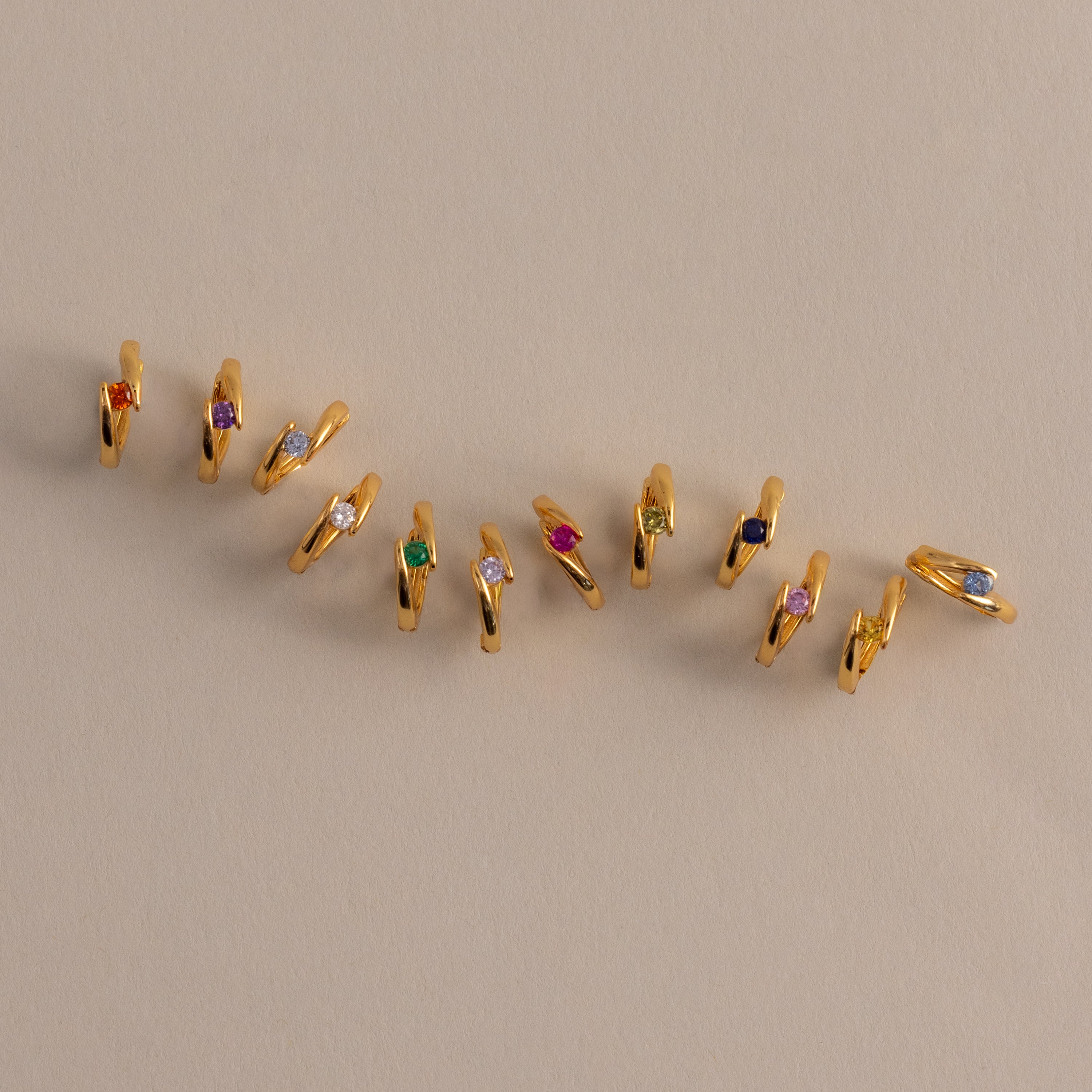 Floating Birthstone Huggies—gold hoops with vibrant gemstones—are arranged diagonally on a beige surface, ideal for fans of personalized jewelry.