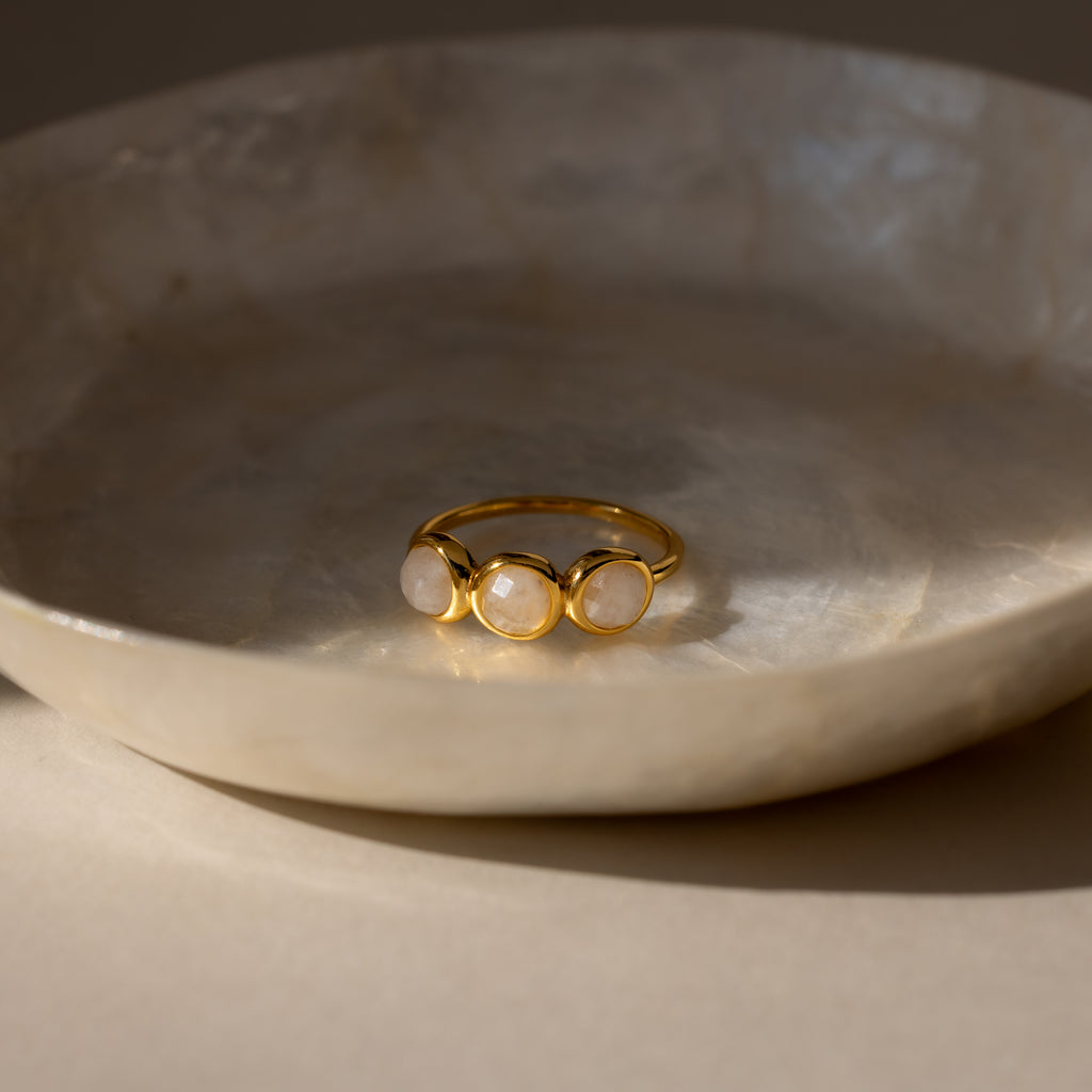 The Moonstone Cluster Ring with three round stones rests in a textured, cream-colored dish under soft lighting.
