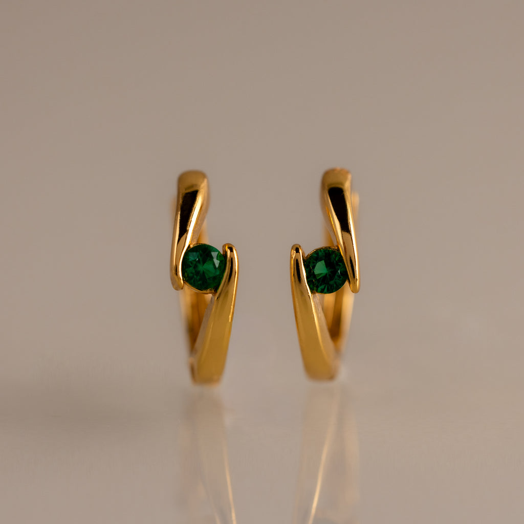 Floating Emerald Huggies, featuring gold hoops with green emerald-like stones, stand upright on a smooth, neutral background.