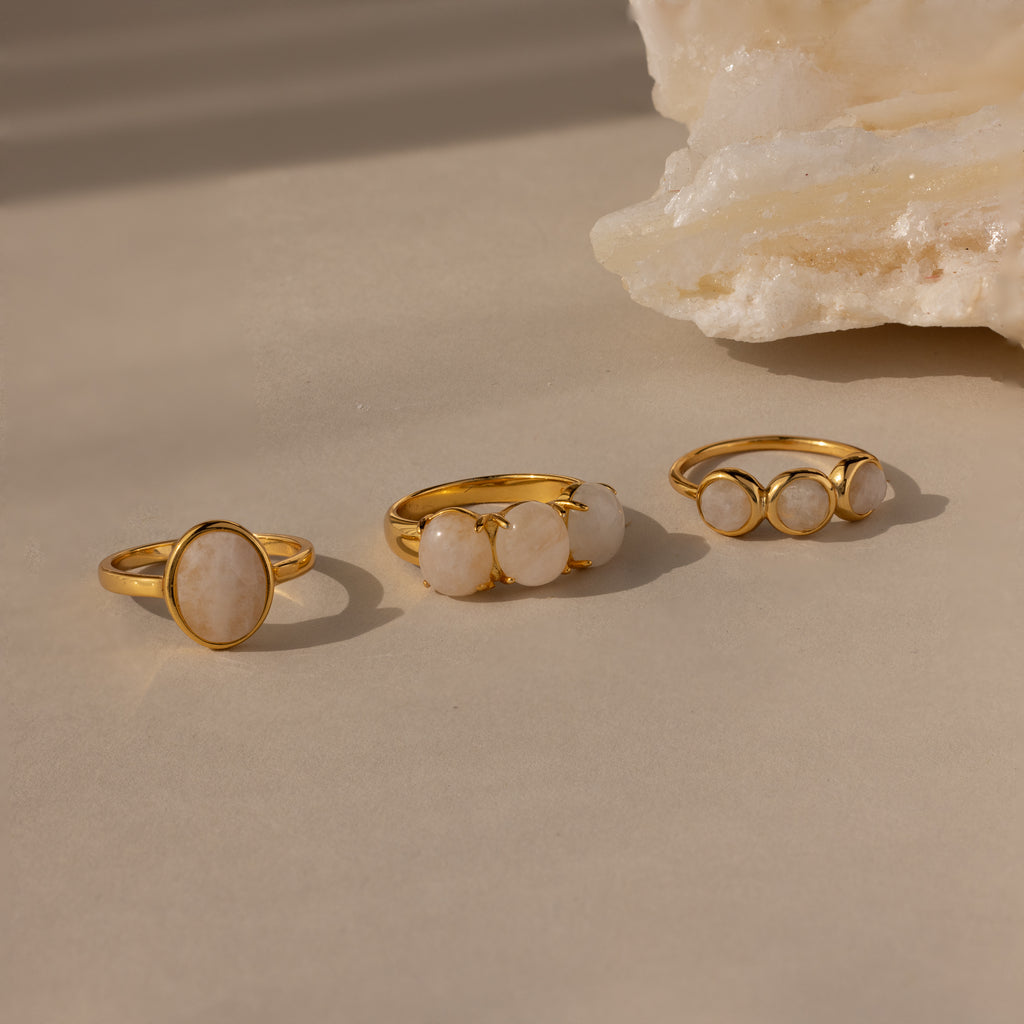 The Moonstone Cluster Ring, a modern gold ring featuring white gemstones, is displayed on a beige surface next to a light-colored stone.