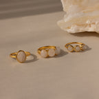 The Moonstone Cluster Ring, a modern gold ring featuring white gemstones, is displayed on a beige surface next to a light-colored stone.