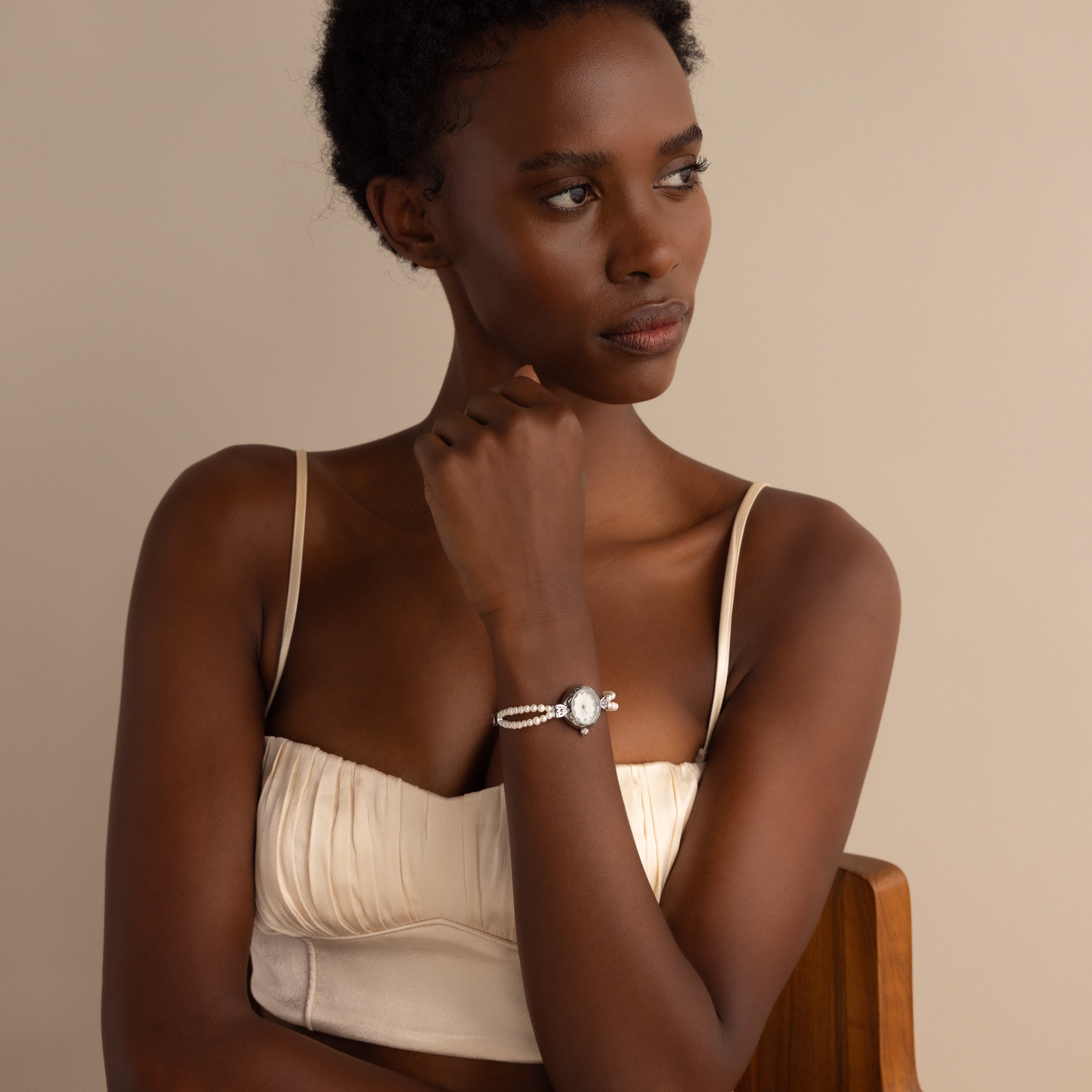 A woman in a cream top sits on a chair, thoughtfully gazing to the side while wearing the Beaded Pearl Timepiece in Sterling Silver.