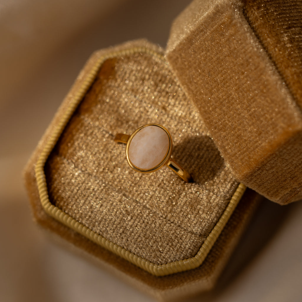 The Moonstone Signet Ring showcases an oval white moonstone on a gold minimalist band, elegantly displayed in an open textured beige velvet box—a perfect choice for everyday jewelry style.