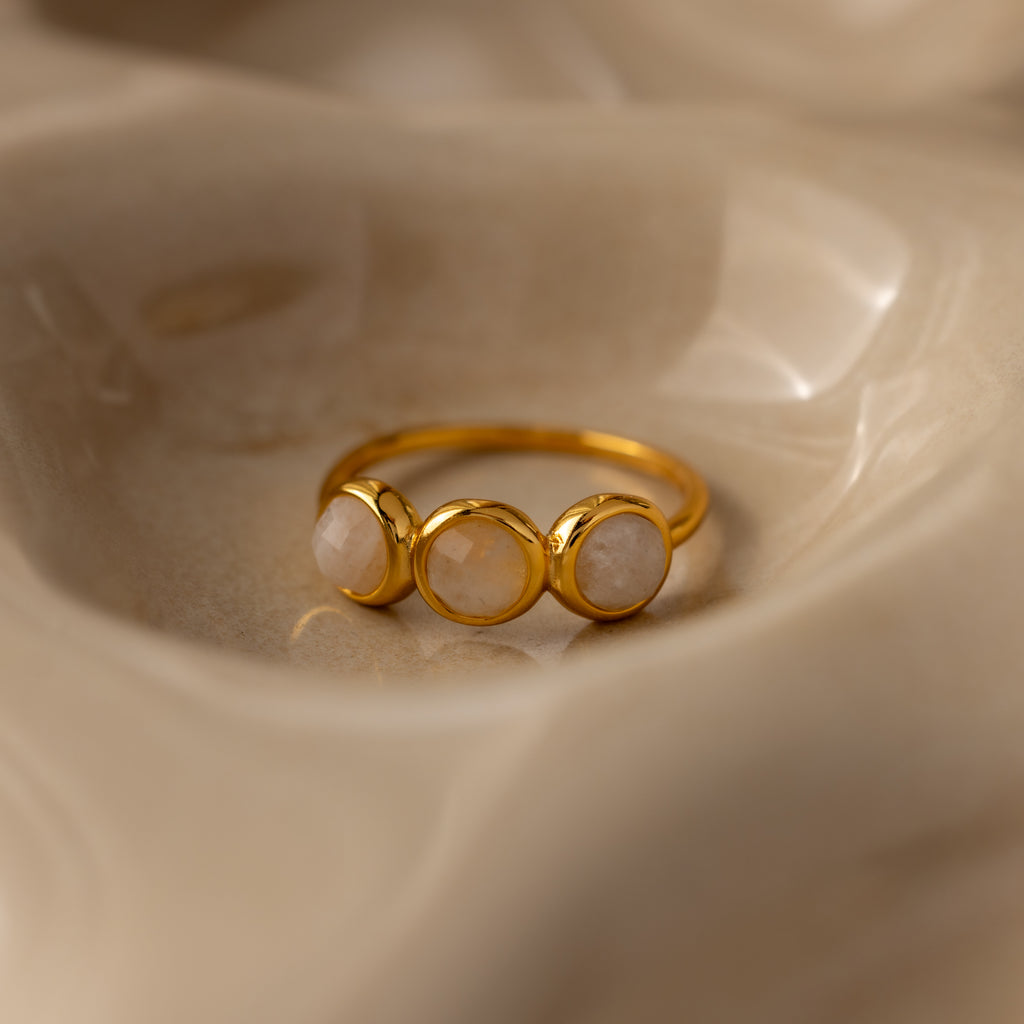The Moonstone Cluster Ring, featuring three round pale moonstones, rests on a smooth beige surface—an elegant example of modern jewelry.