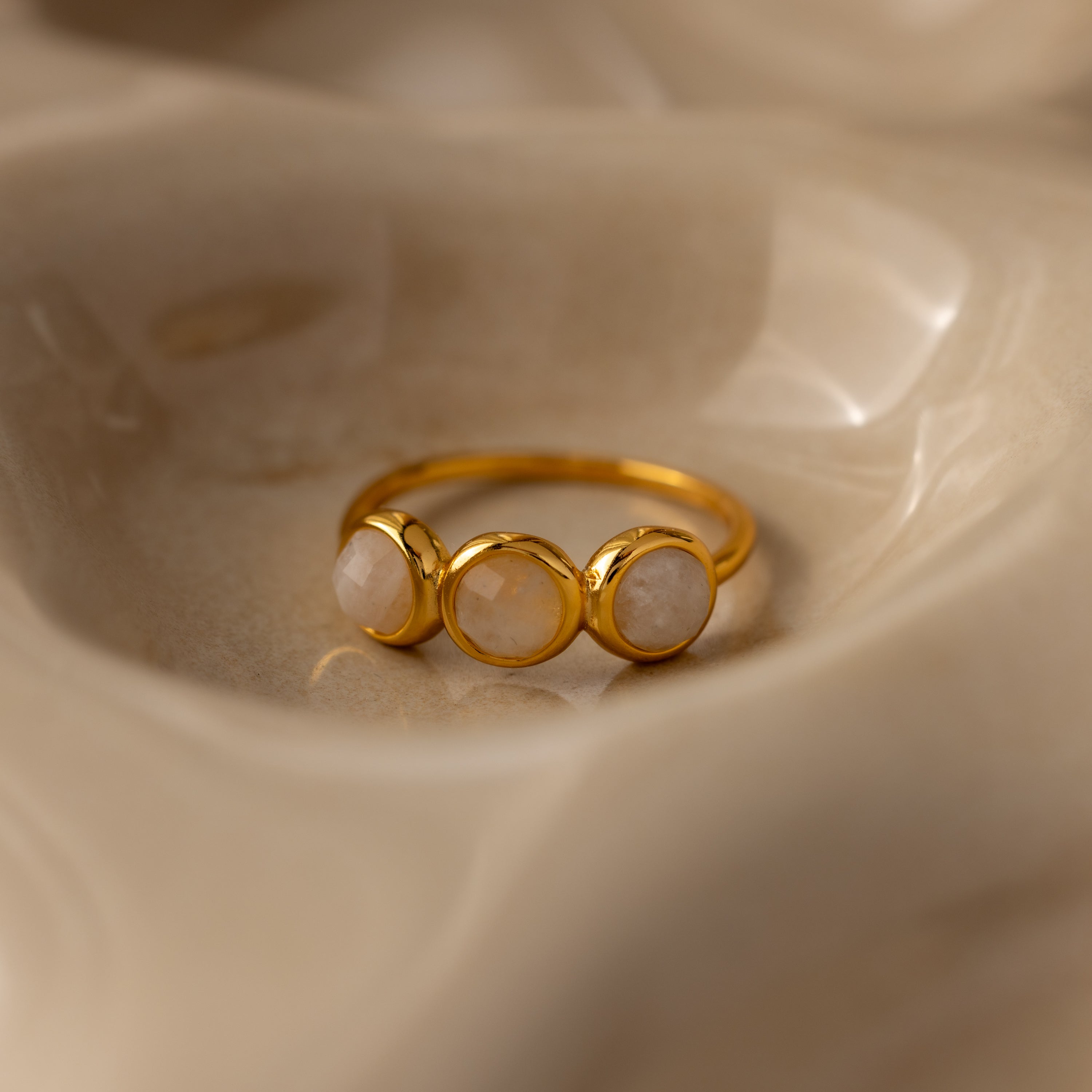 The Moonstone Cluster Ring, featuring three round pale moonstones, rests on a smooth beige surface—an elegant example of modern jewelry.
