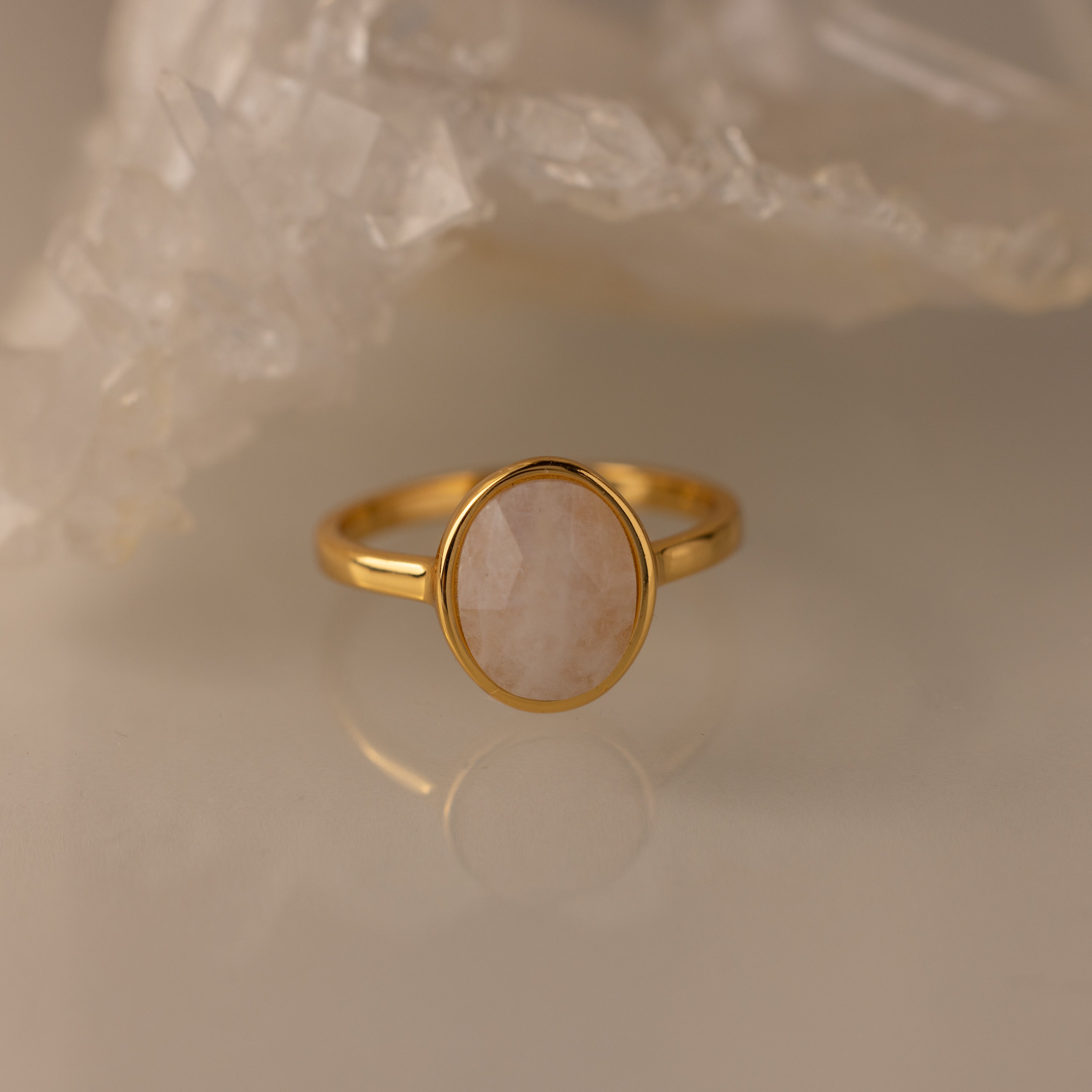 The Moonstone Signet Ring, featuring a gold minimalist band and moonstone gemstone, rests on a glossy surface near a cluster of crystals—an ideal choice for fans of everyday jewelry.