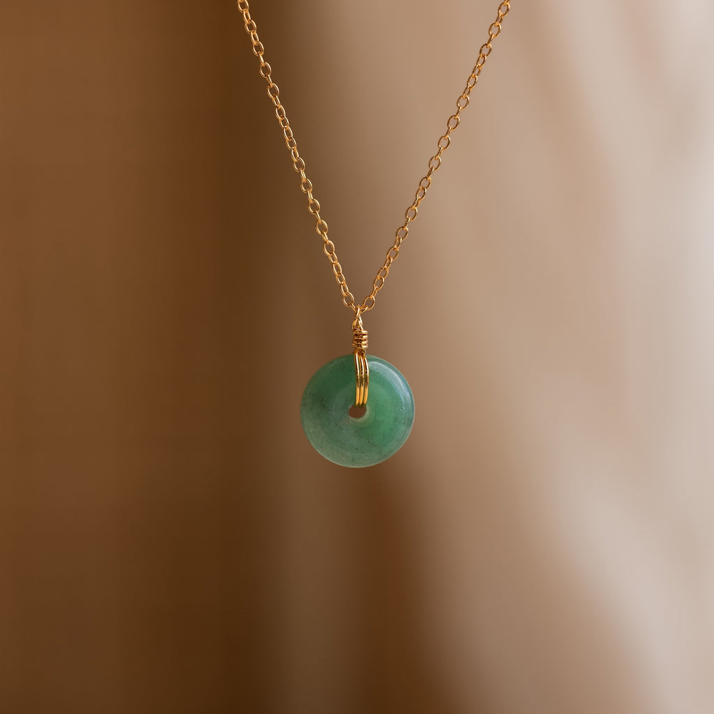 The Round Jade Pendant Necklace features a gold chain with a round jade gemstone, displayed on a soft beige background for a naturally elegant look.