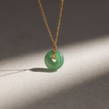 The Round Jade Pendant Necklace features a green jade gemstone on a gold chain, displayed on a light stone surface with soft shadows—highlighting natural elegance.