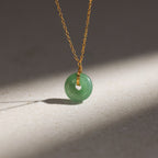 The Round Jade Pendant Necklace features a green jade gemstone on a gold chain, displayed on a light stone surface with soft shadows—highlighting natural elegance.