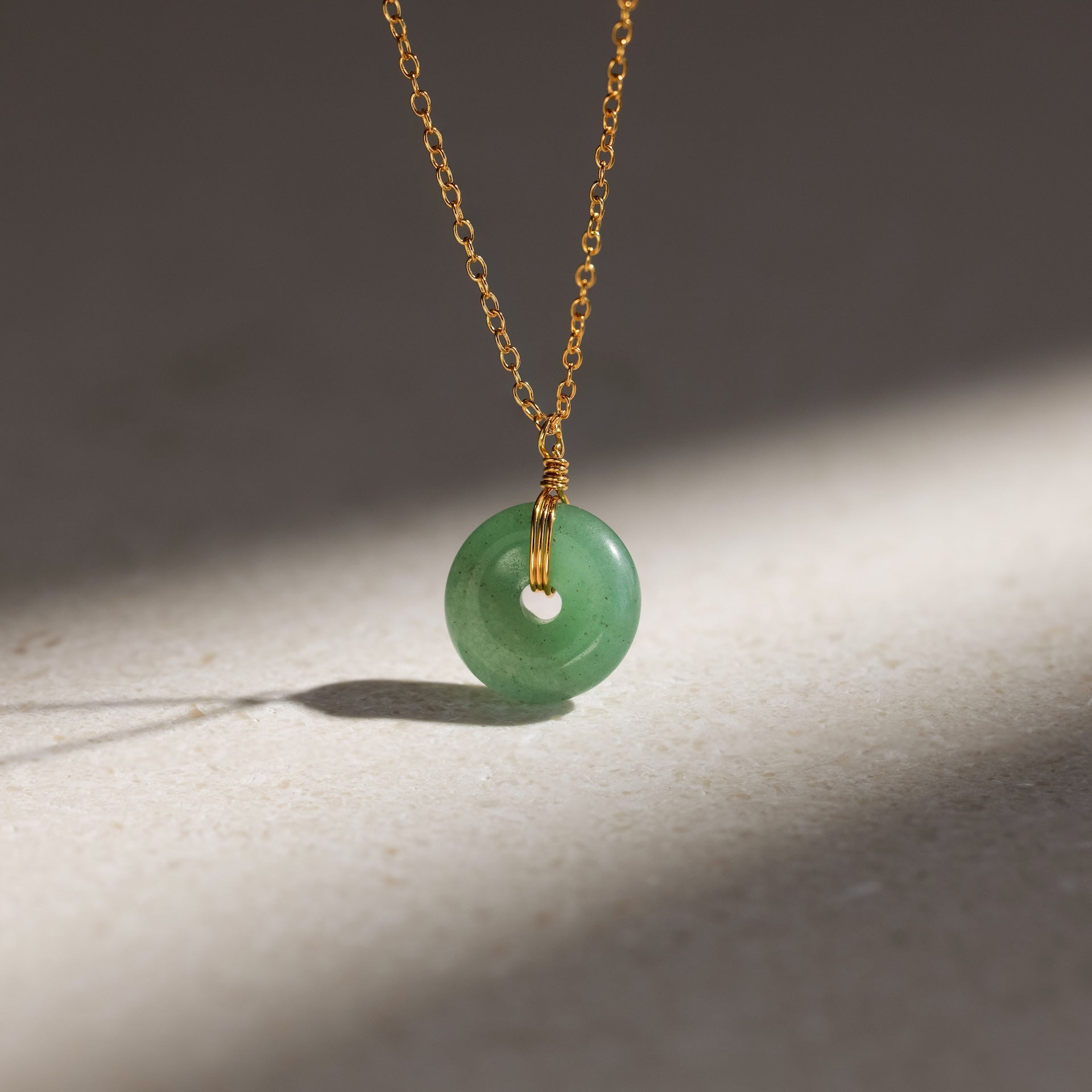 The Round Jade Pendant Necklace features a green jade gemstone on a gold chain, displayed on a light stone surface with soft shadows—highlighting natural elegance.