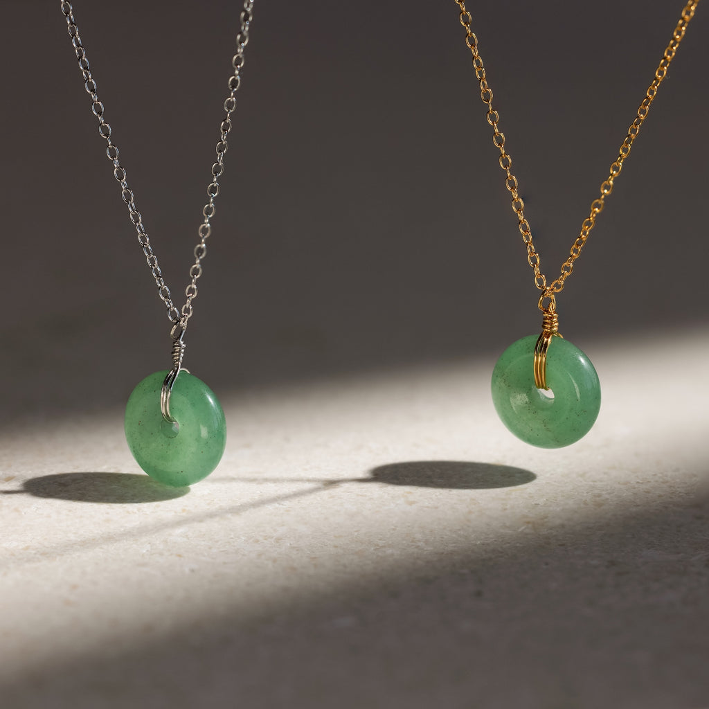 A Round Jade Pendant Necklace in 18K Gold casts an elegant shadow as its jade gemstone hangs gracefully from a gold chain on a light surface.