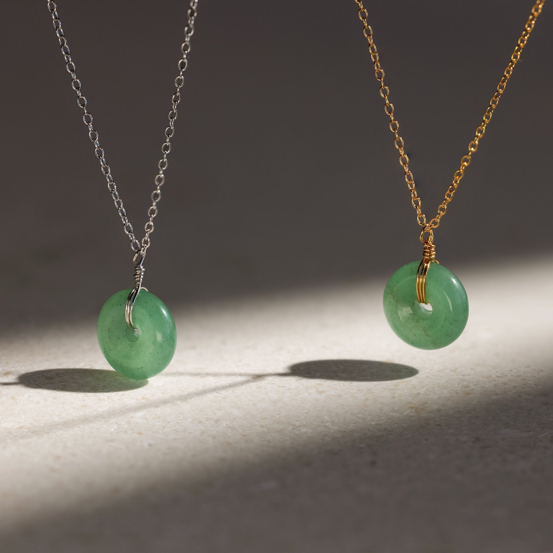 A Round Jade Pendant Necklace in 18K Gold casts an elegant shadow as its jade gemstone hangs gracefully from a gold chain on a light surface.