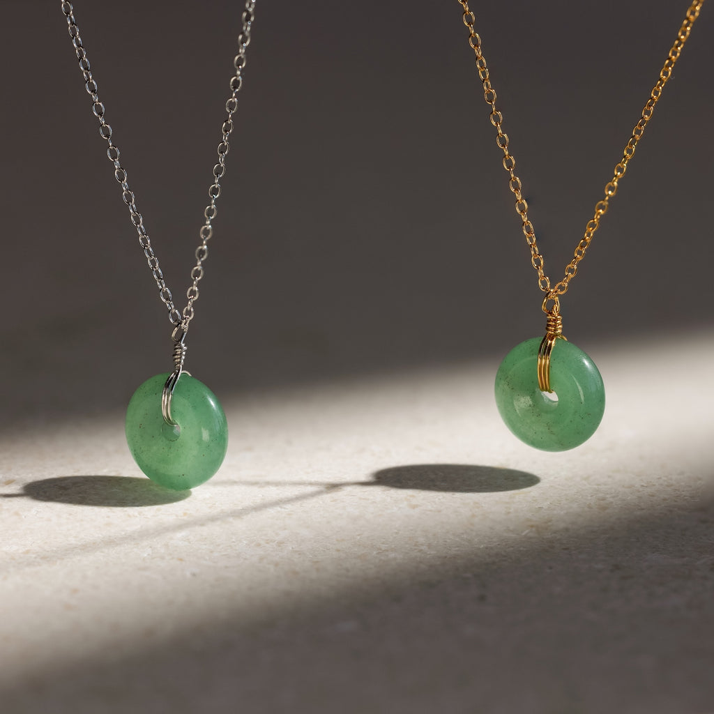 Two Round Jade Pendant Necklaces—one on a silver chain, one on gold—feature green gemstone pendants and cast elegant shadows on a light surface.