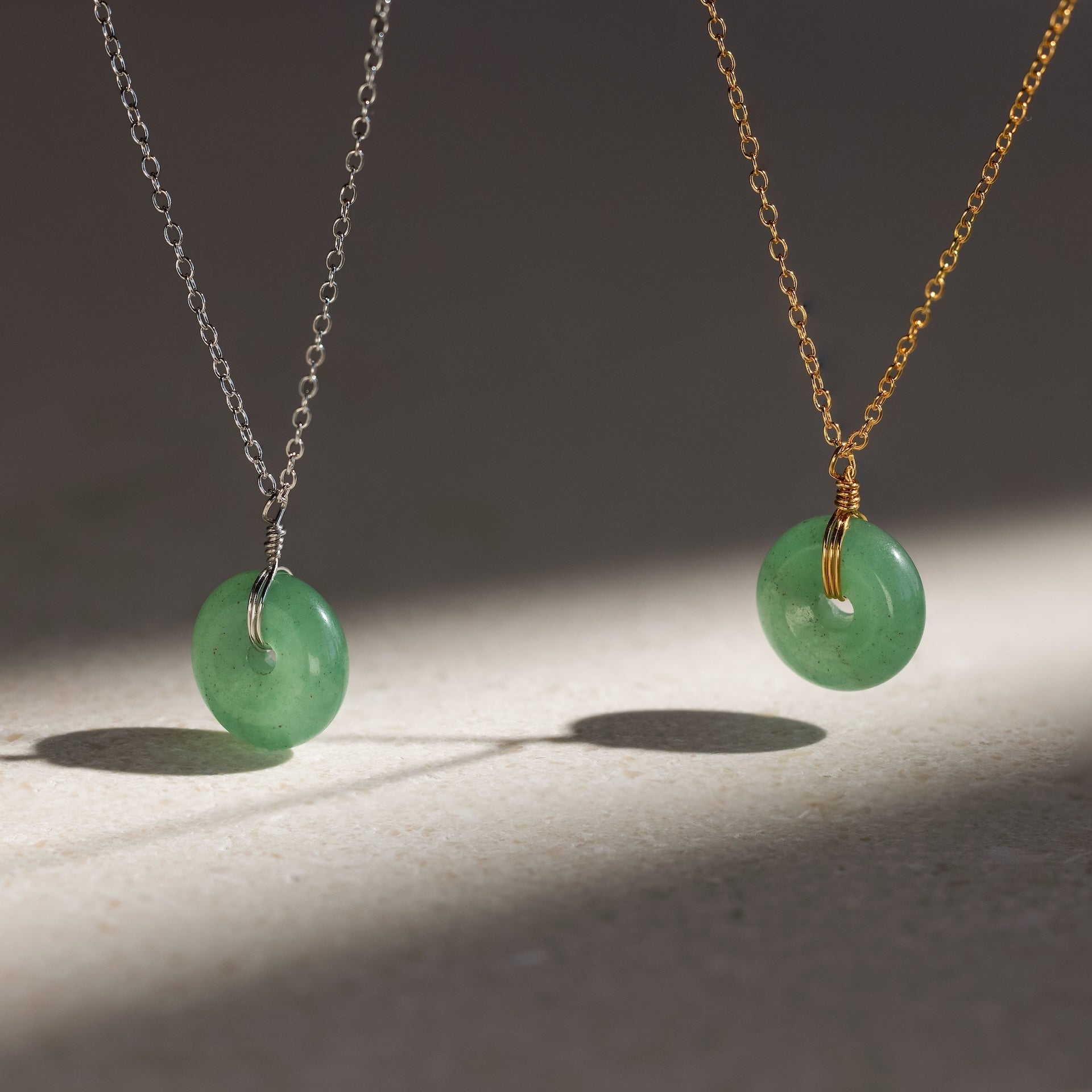 Two Round Jade Pendant Necklaces—one on a silver chain, one on gold—feature green gemstone pendants and cast elegant shadows on a light surface.