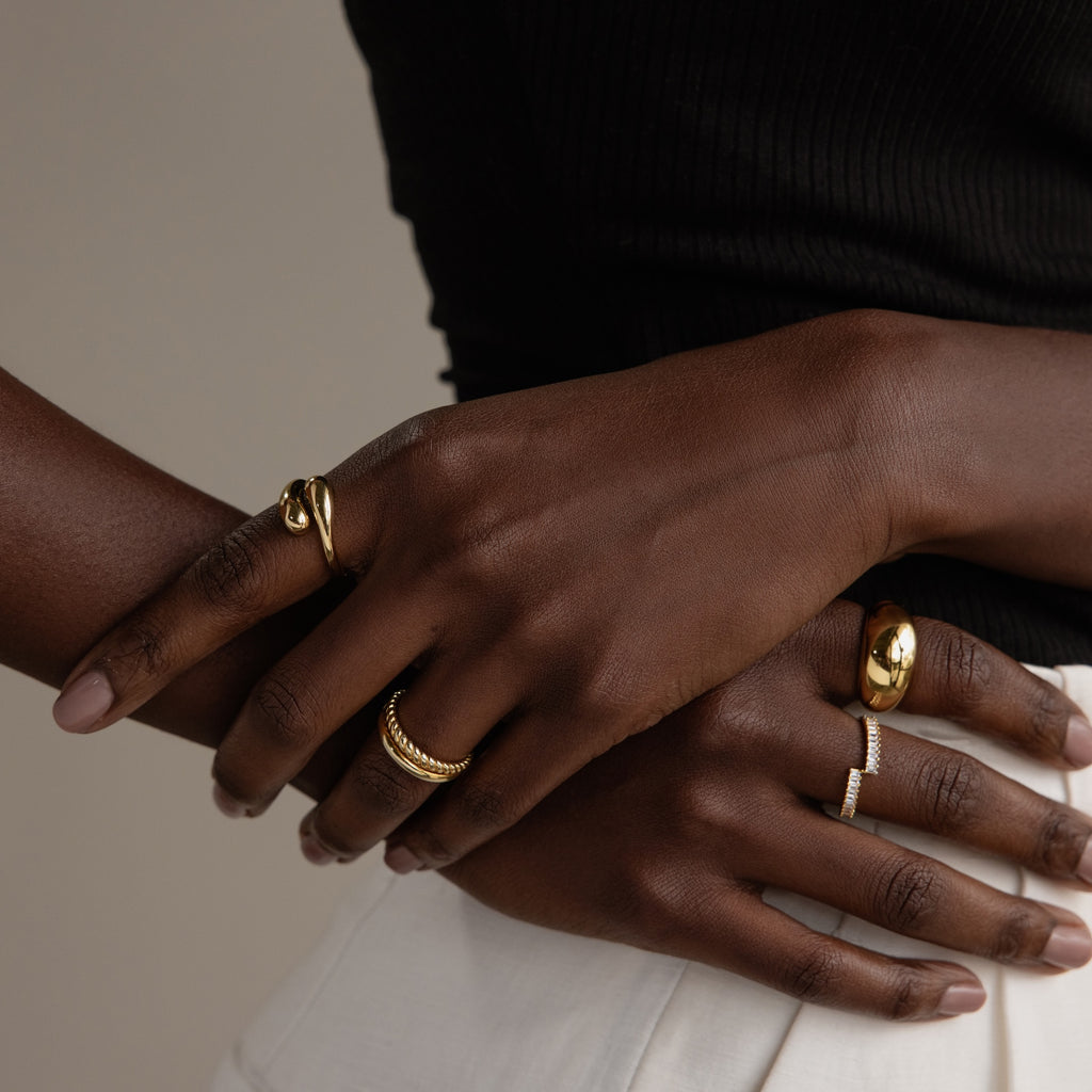 Close-up of crossed hands showcasing sculptural jewelry, including the Duo Dome Ring with an adjustable band, against a neutral backdrop; the person is dressed in a black top and white pants.