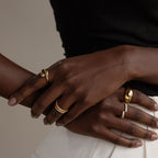 Close-up of crossed hands showcasing sculptural jewelry, including the Duo Dome Ring with an adjustable band, against a neutral backdrop; the person is dressed in a black top and white pants.