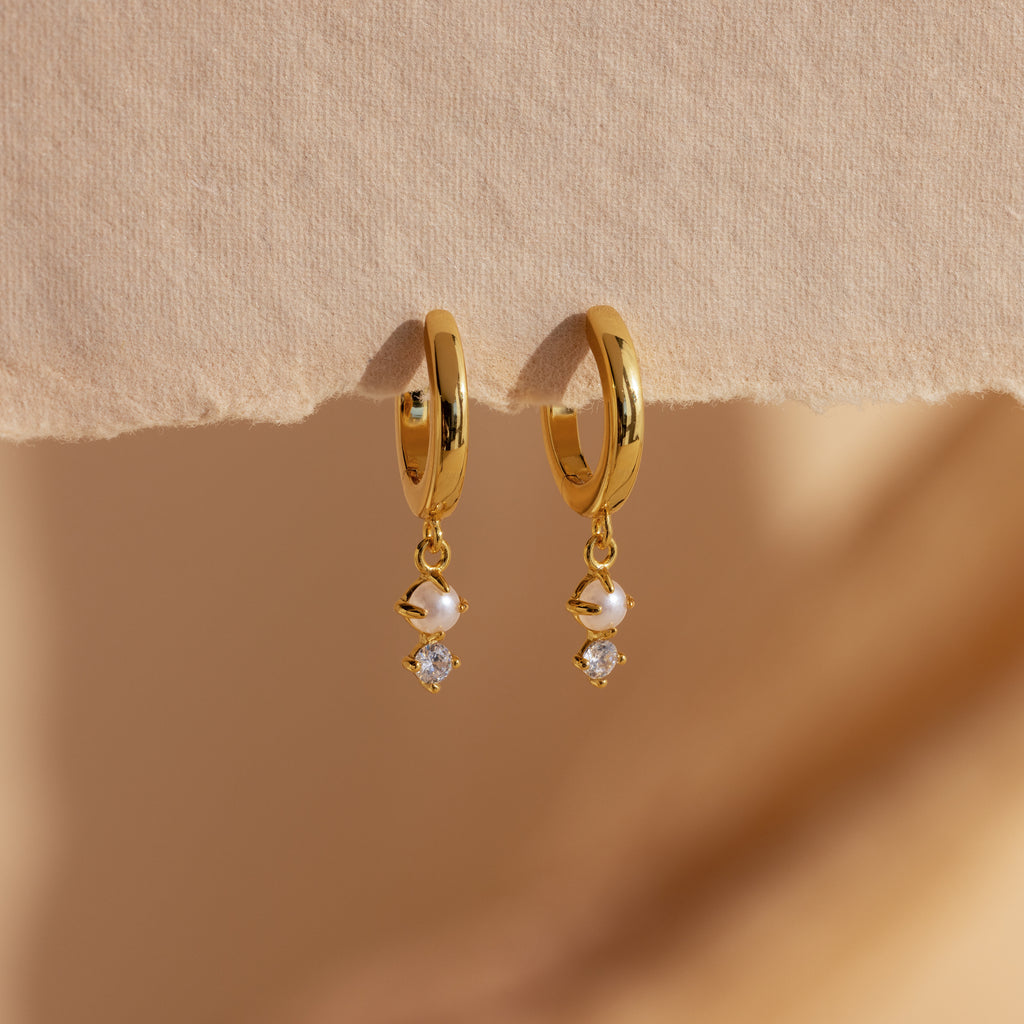 Dangling Pearl Diamond Huggies in 18K Gold, featuring elegant hoops with pearls and sparkling diamonds, are displayed on a textured beige surface.