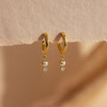 Dangling Pearl Diamond Huggies in 18K Gold, featuring elegant hoops with pearls and sparkling diamonds, are displayed on a textured beige surface.