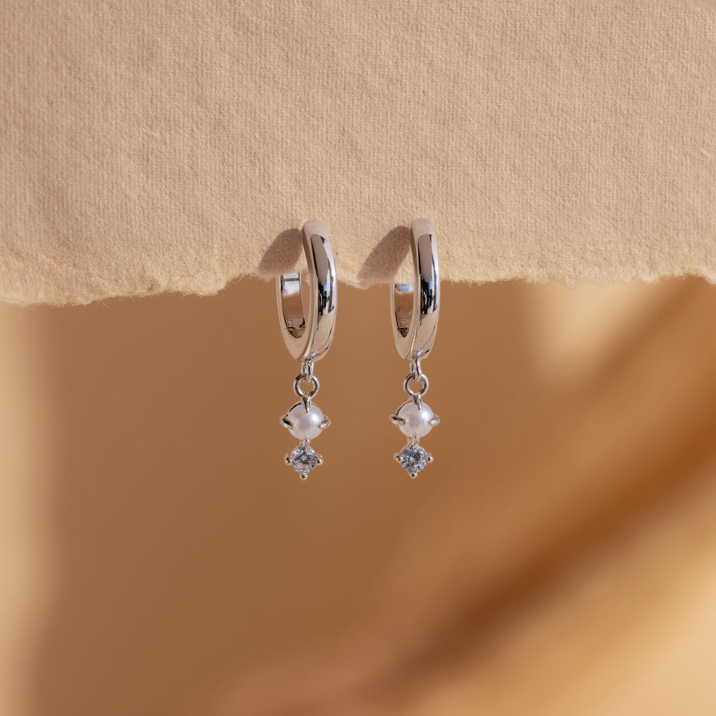 The Dangling Pearl Diamond Huggies in Sterling Silver shimmer with pearl and diamond-like dangles, elegantly displayed against a beige textured background.