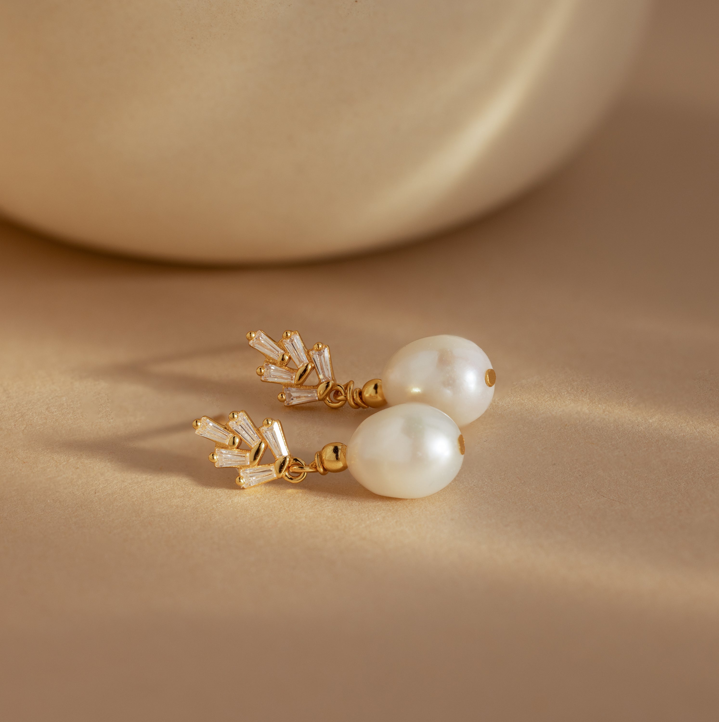 Pearl Diamond Drop Earrings with crystal accents rest on a beige surface, softly lit—vintage-inspired earrings for an elegant touch.