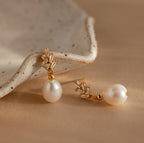 Pearl Diamond Drop Earrings in 18K Gold with crystal accents, vintage-inspired and elegant, are displayed on a beige surface next to a ceramic dish.