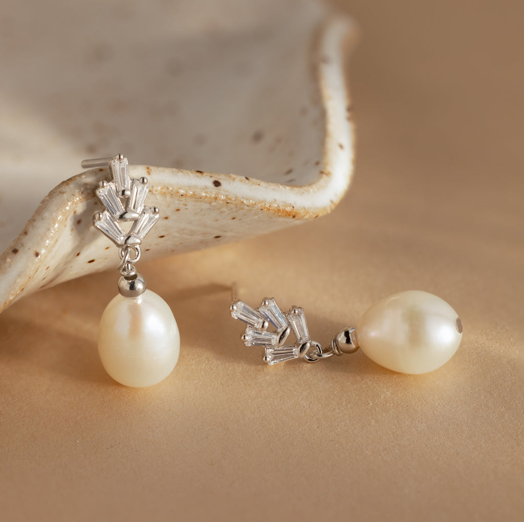 The Pearl Diamond Drop Earrings, featuring vintage-inspired pearl drops and silver accents, are displayed on a beige surface next to a ceramic dish.