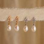 Two pairs of Pearl Diamond Drop Earrings—one with gold and one with silver accents—are displayed elegantly on soft beige fabric.