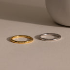 The Minimal Stacking Ring in gold and silver rests side by side on a beige surface, with a white round object nearby.