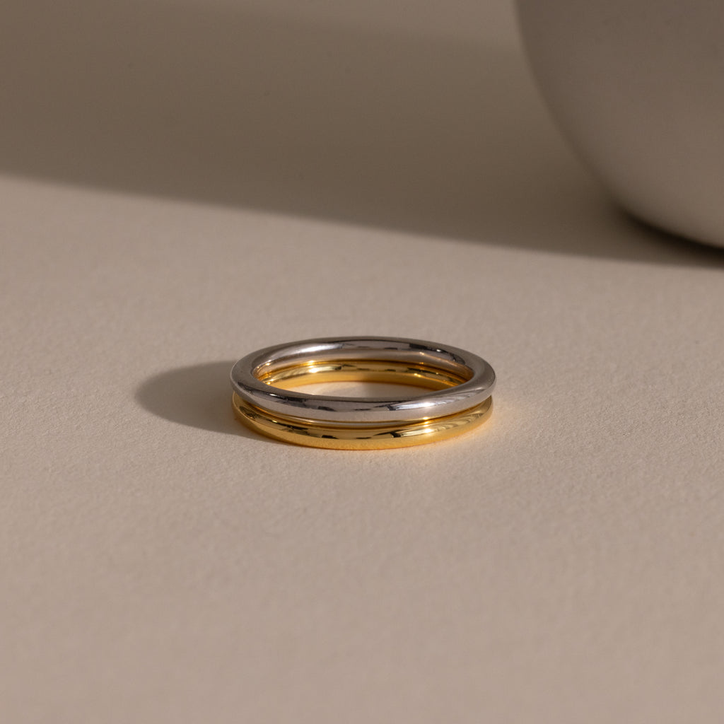 The Minimal Stacking Ring, featuring a silver and gold two-tone design, lies on a beige surface with a soft shadow next to a round object.