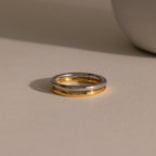 The Minimal Stacking Ring, featuring a silver and gold two-tone design, lies on a beige surface with a soft shadow next to a round object.