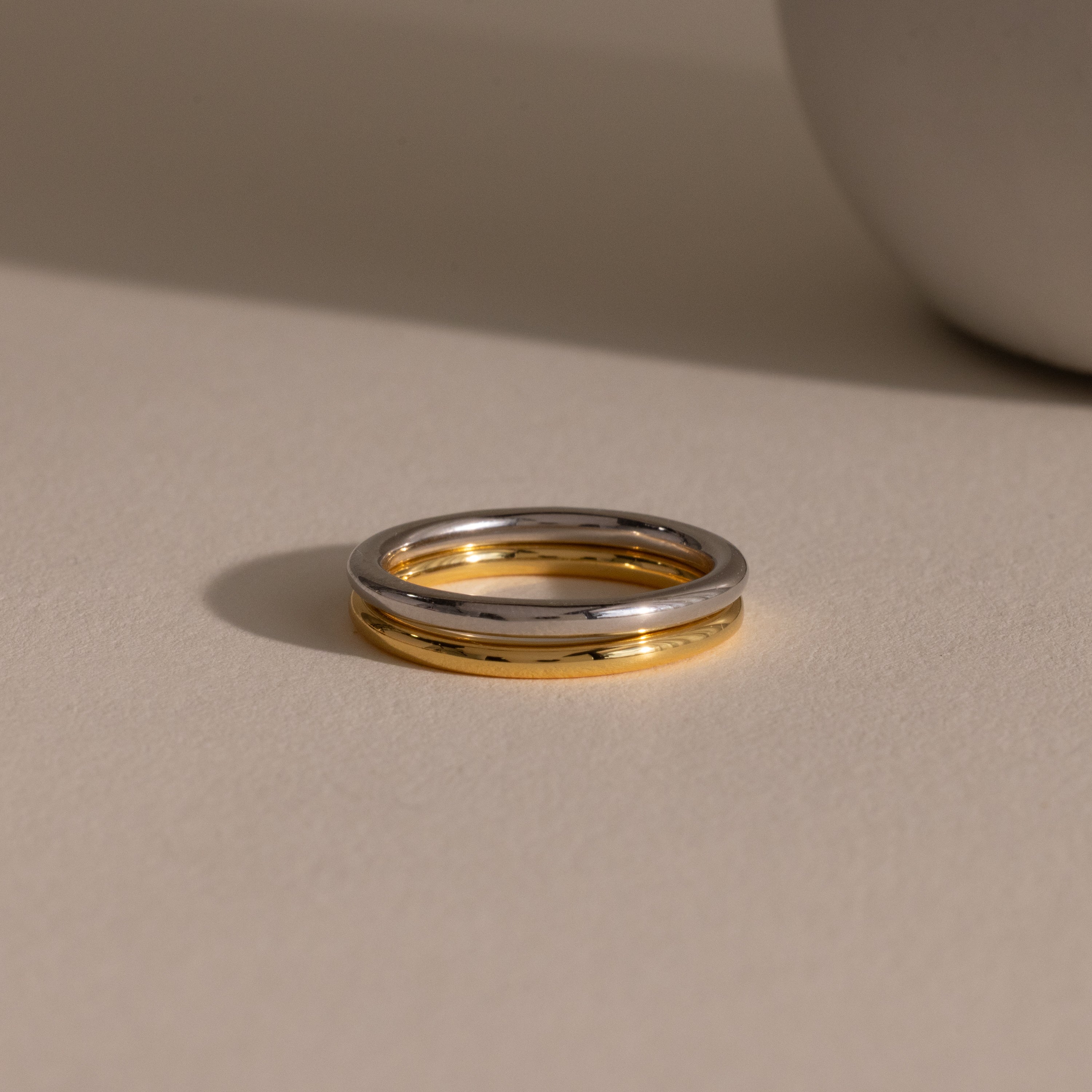 The Minimal Stacking Ring, featuring a silver and gold two-tone design, lies on a beige surface with a soft shadow next to a round object.