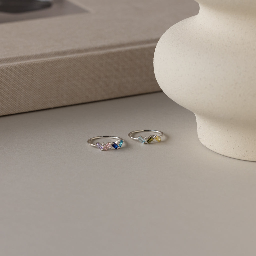 The Cluster Baguette Birthstone Rings in Sterling Silver, featuring colorful gemstones, are displayed on a neutral surface near a textured vase and a closed book—a perfect personalized statement piece.