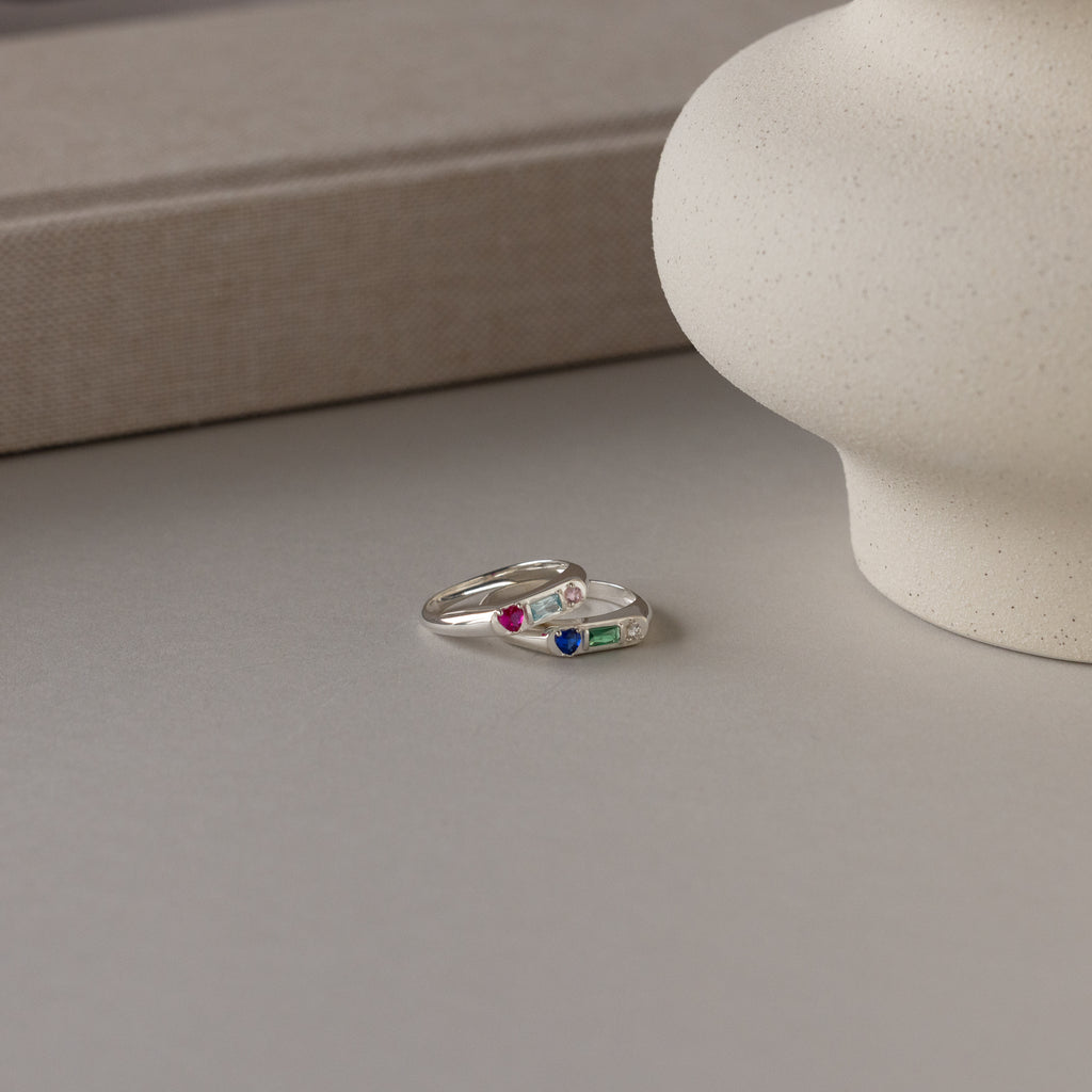 The Bold Birthstone Signet Ring in Sterling Silver with colorful gemstones rests on a neutral surface alongside another silver ring and a round ceramic vase.