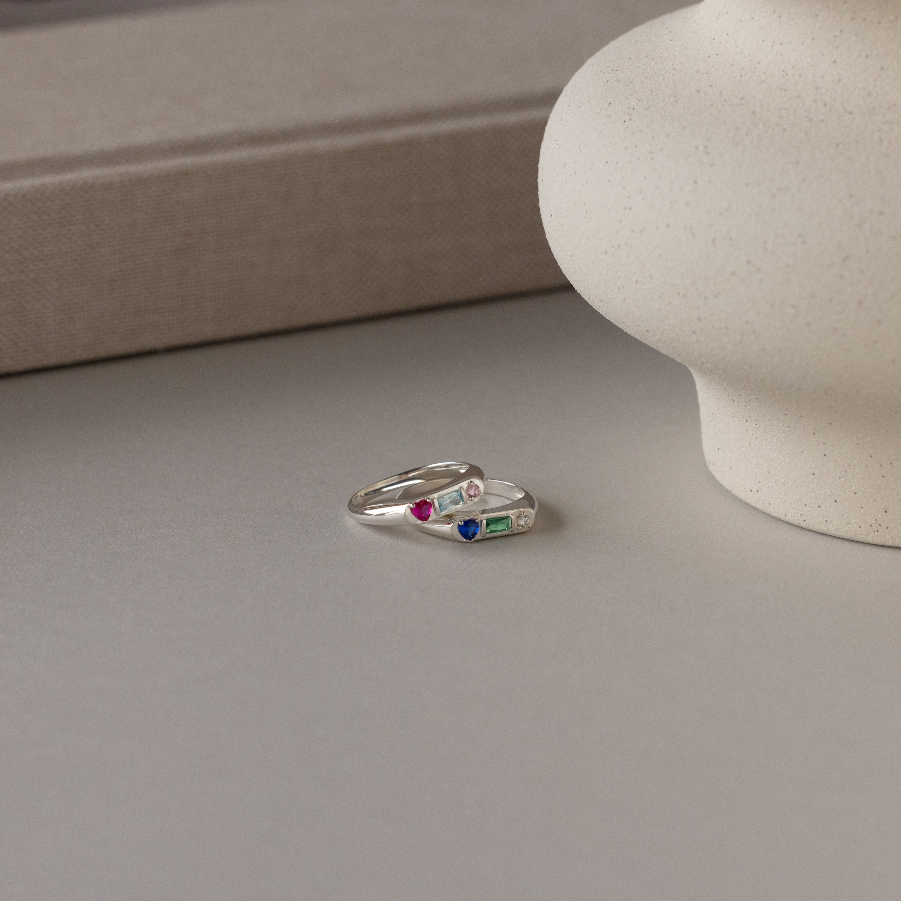 The Bold Birthstone Signet Ring in Sterling Silver with colorful gemstones rests on a neutral surface alongside another silver ring and a round ceramic vase.