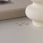 Two Alex Initial Birthstone Rings in sterling silver, featuring letter charms and small gemstones, are displayed on a neutral surface next to a textured vase and a book.