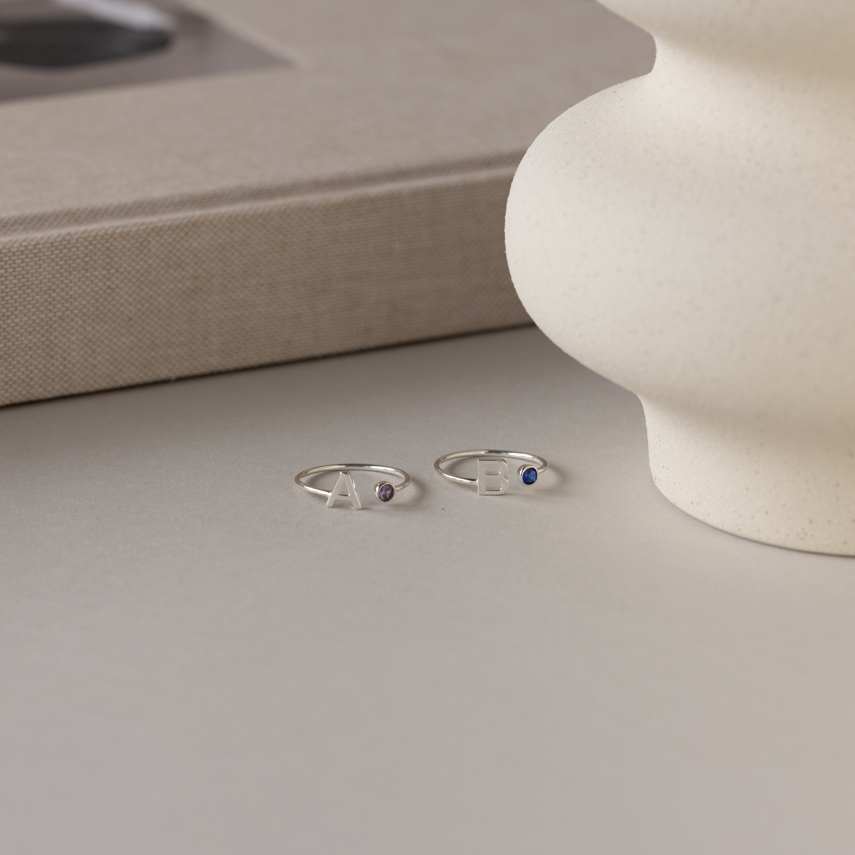 Two Alex Initial Birthstone Rings in sterling silver, featuring letter charms and small gemstones, are displayed on a neutral surface next to a textured vase and a book.
