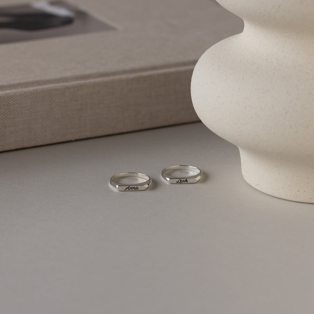The Paris Thin Name Signet Ring in Sterling Silver rests on a light surface beside a textured vase and a closed book, exuding minimalist charm.