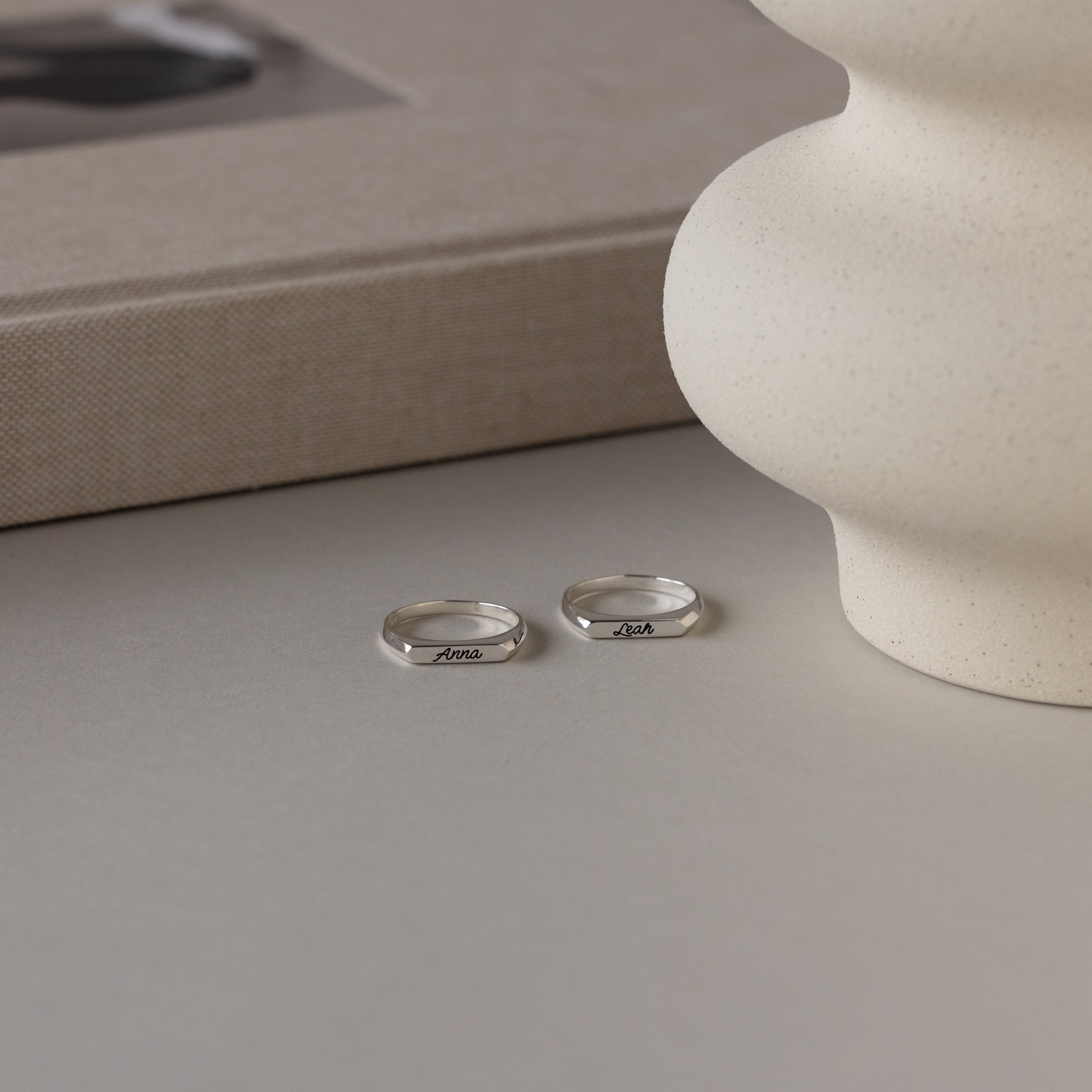 The Paris Thin Name Signet Ring in Sterling Silver rests on a light surface beside a textured vase and a closed book, exuding minimalist charm.