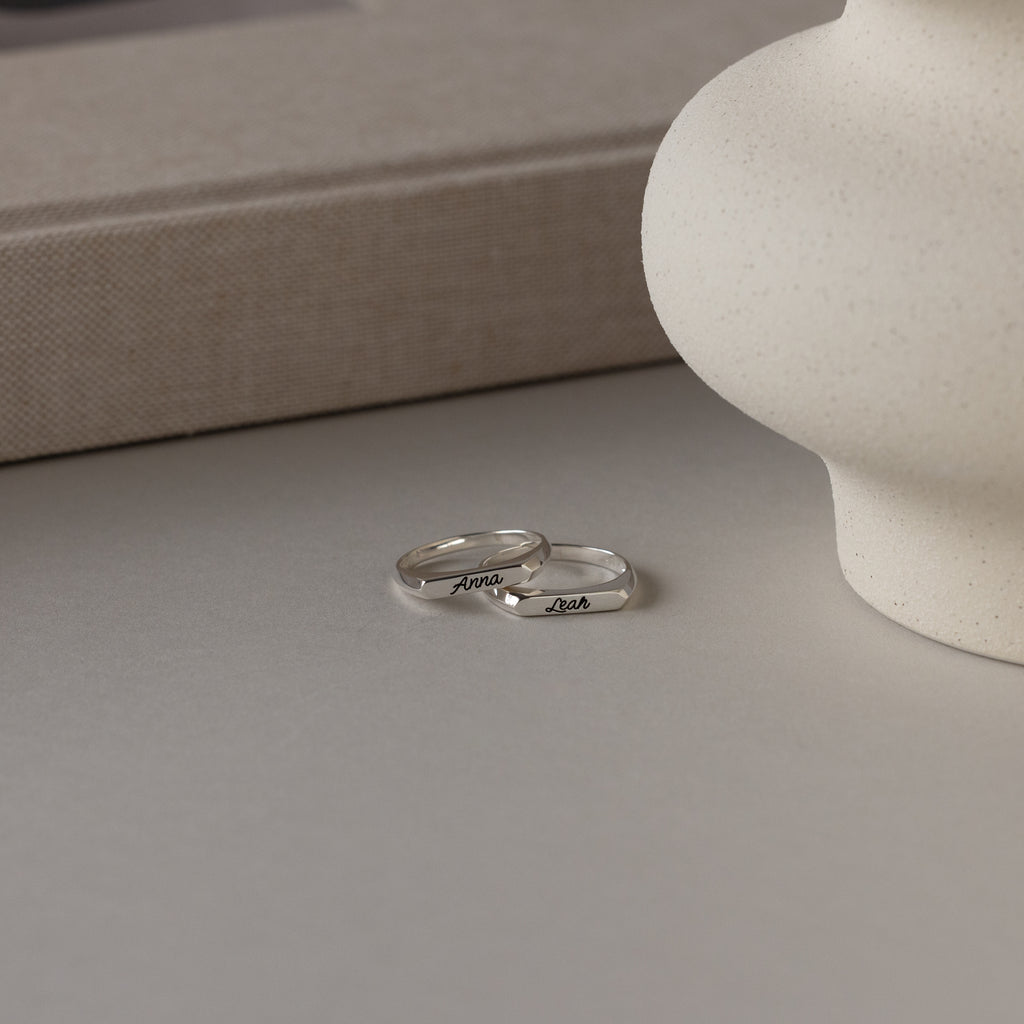 A Paris Thin Name Signet Ring in Sterling Silver, custom-engraved with the names Anna and Josh, rests on a neutral-colored surface beside a round vase.