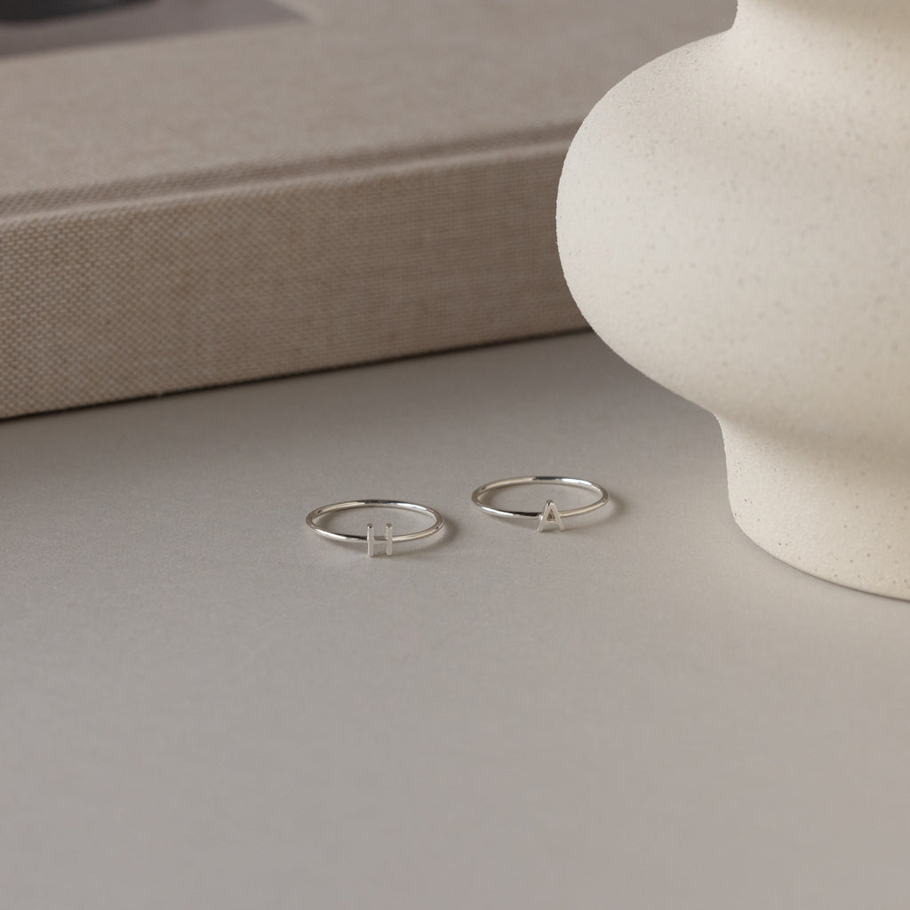 The Alex Dainty Letter Ring in Sterling Silver features two minimalist rings with small H and A letter charms, resting on a neutral surface near a white vase—ideal as personalized or dainty everyday jewelry.