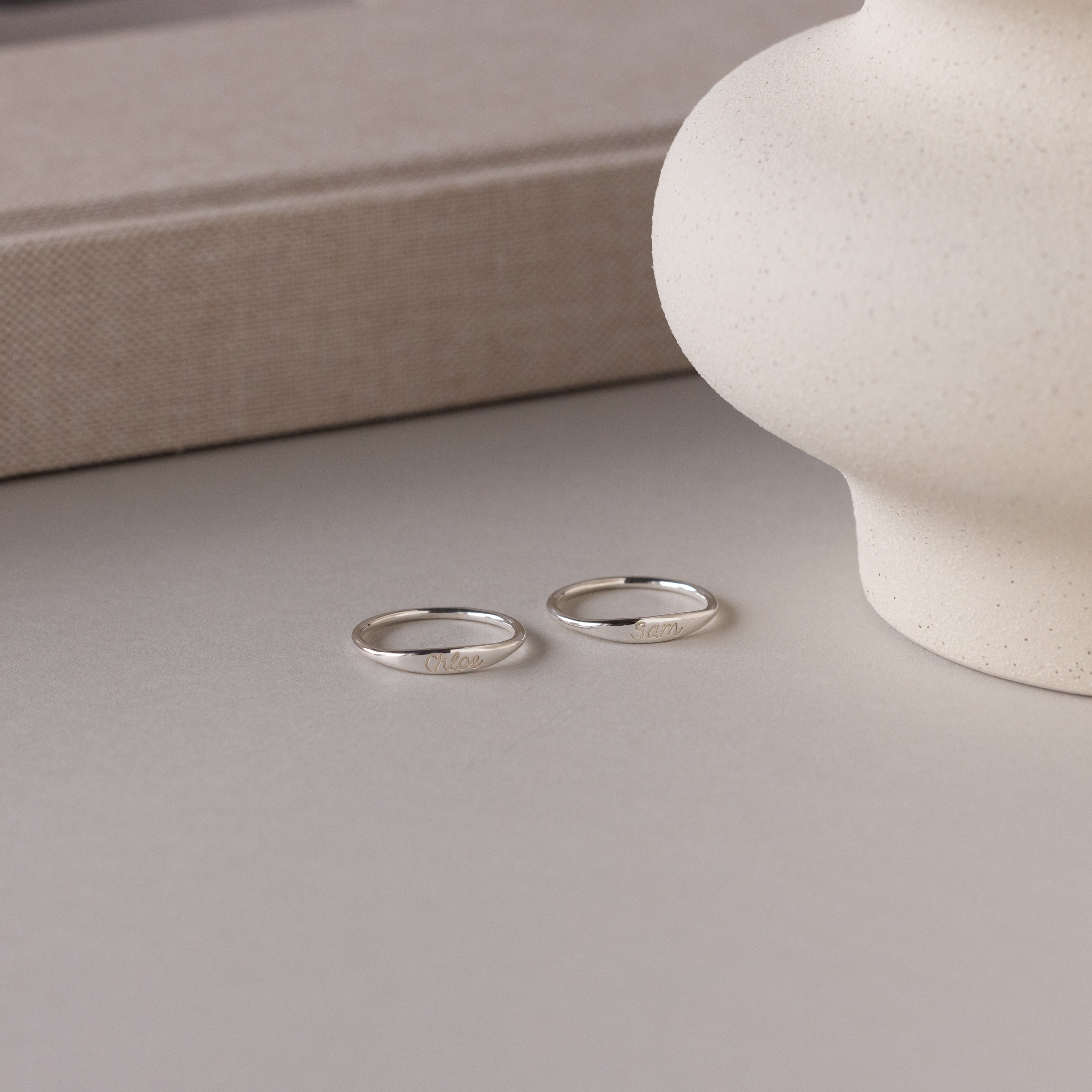 The Ann Skinny Name Ring in Sterling Silver rests on a light surface beside a textured vase and beige book—an elegant Christmas gift for her.