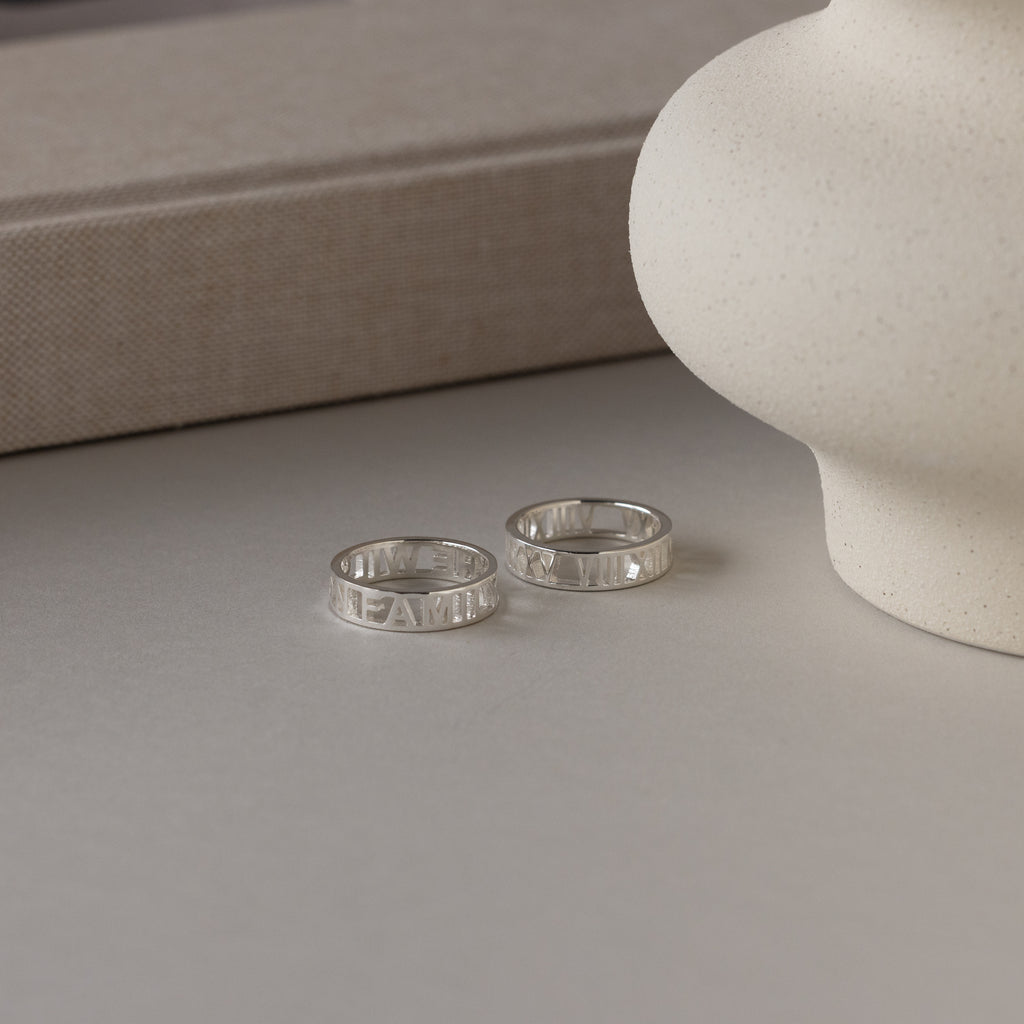Two Roman Numerals Rings in Sterling Silver rest on a light surface beside a white ceramic vase and a beige book—an elegant milestone gift.