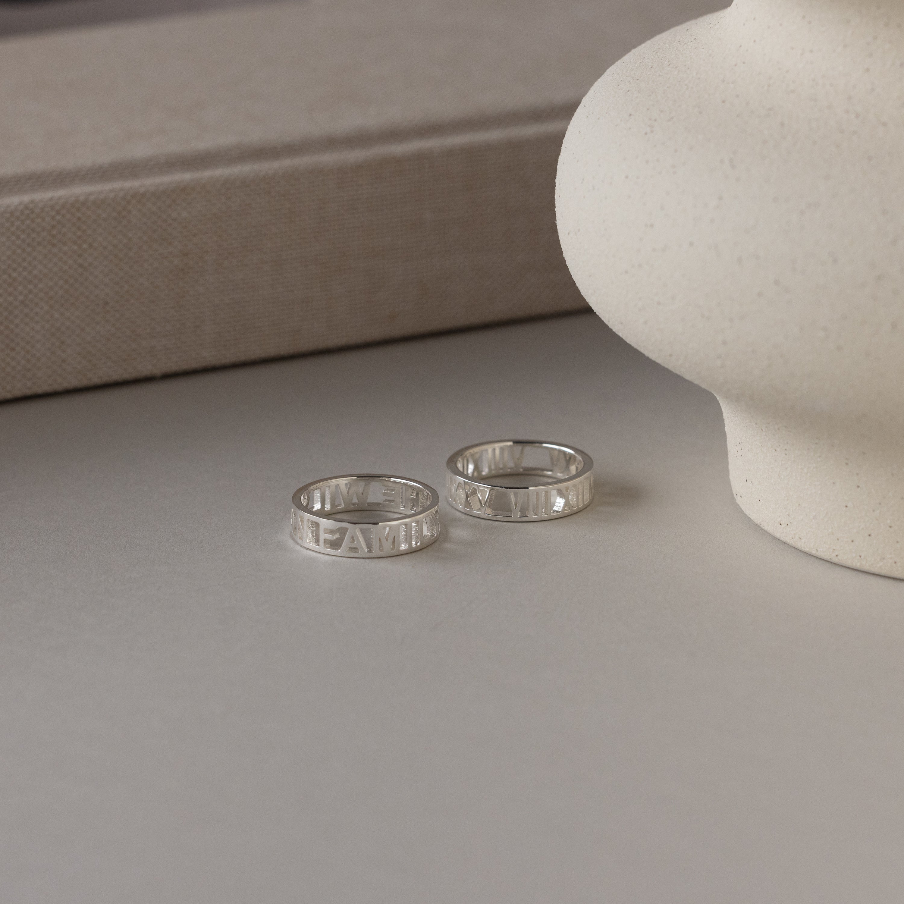 Two Roman Numerals Rings in Sterling Silver rest on a light surface beside a white ceramic vase and a beige book—an elegant milestone gift.