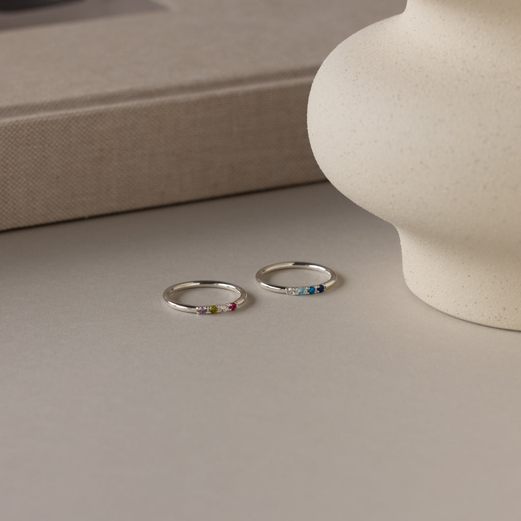 The Mini Birthstone Ring in Sterling Silver, featuring small colorful gemstones, rests on a light surface by a beige vase and book—a perfect personalized piece for stacking or daily wear.