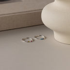 The Mini Birthstone Ring in Sterling Silver, featuring small colorful gemstones, rests on a light surface by a beige vase and book—a perfect personalized piece for stacking or daily wear.