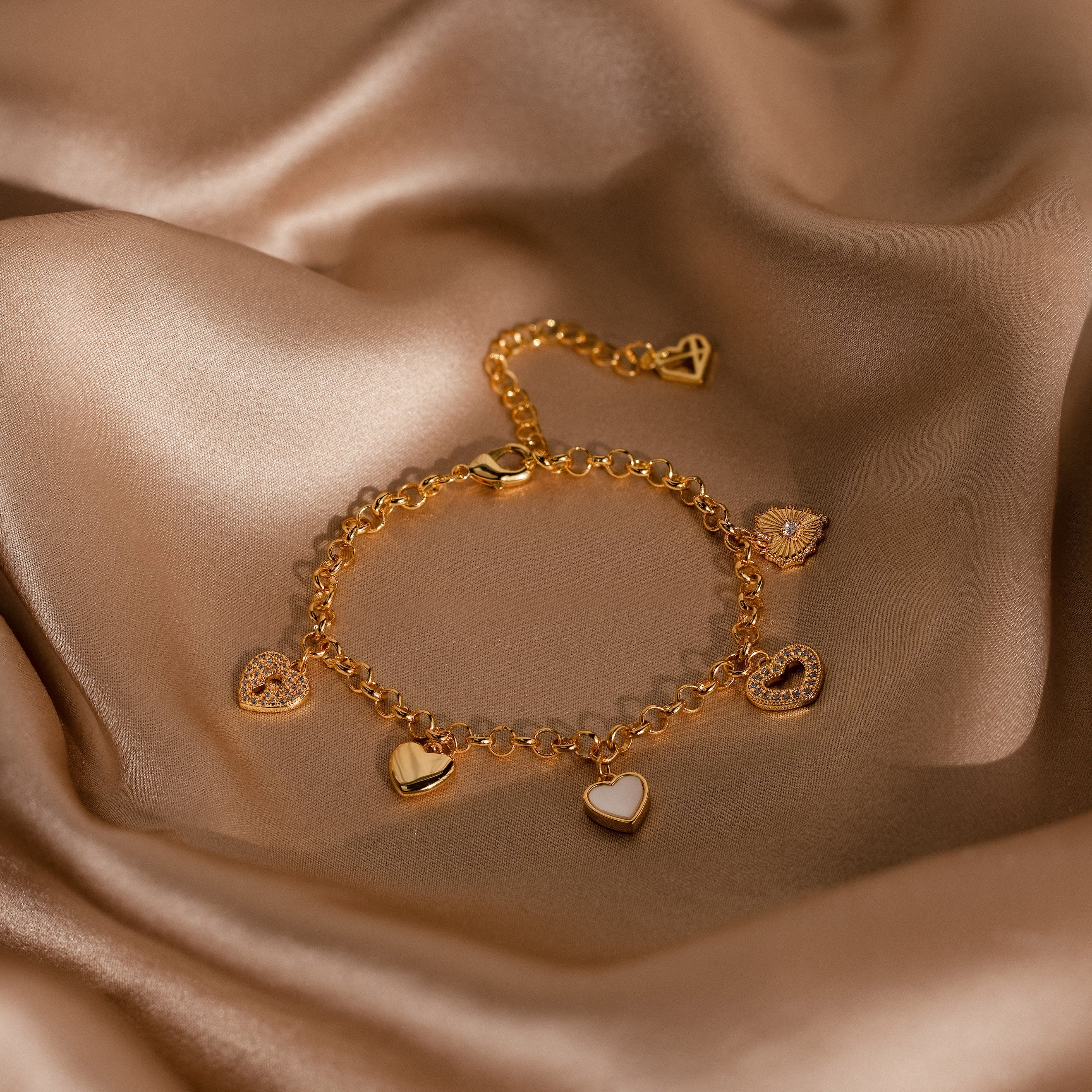The Heart Charm Bracelet in 18K Gold, displayed on smooth champagne satin, features delicate heart charms—an elegant gift for your girlfriend.