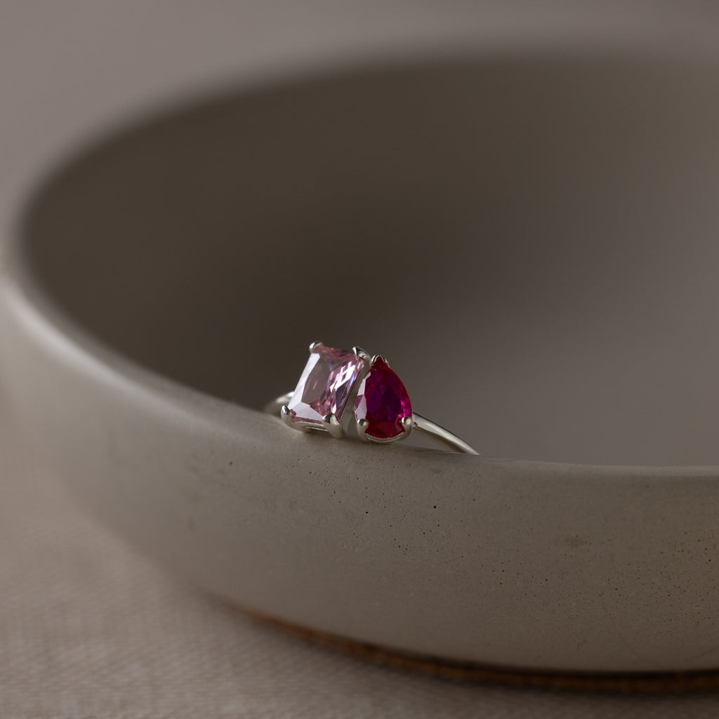The Toi et Moi Birthstone Ring in Sterling Silver, with one pink and one red gemstone, sits on the edge of a round ceramic bowl.